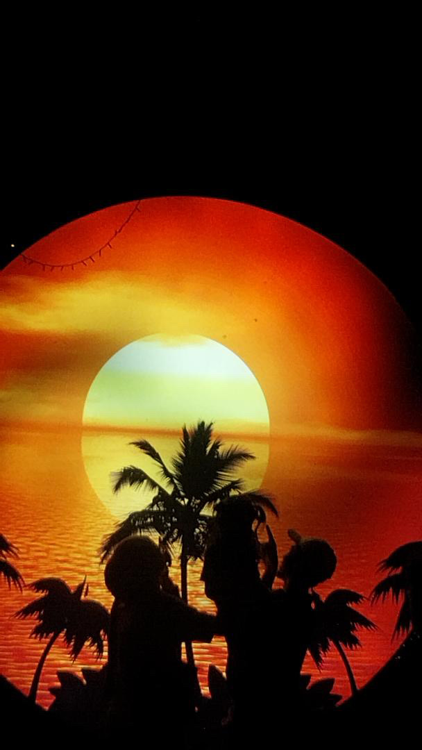 A diorama of a tropical sunset framed in a circular window, palms and figures in silhouette.
