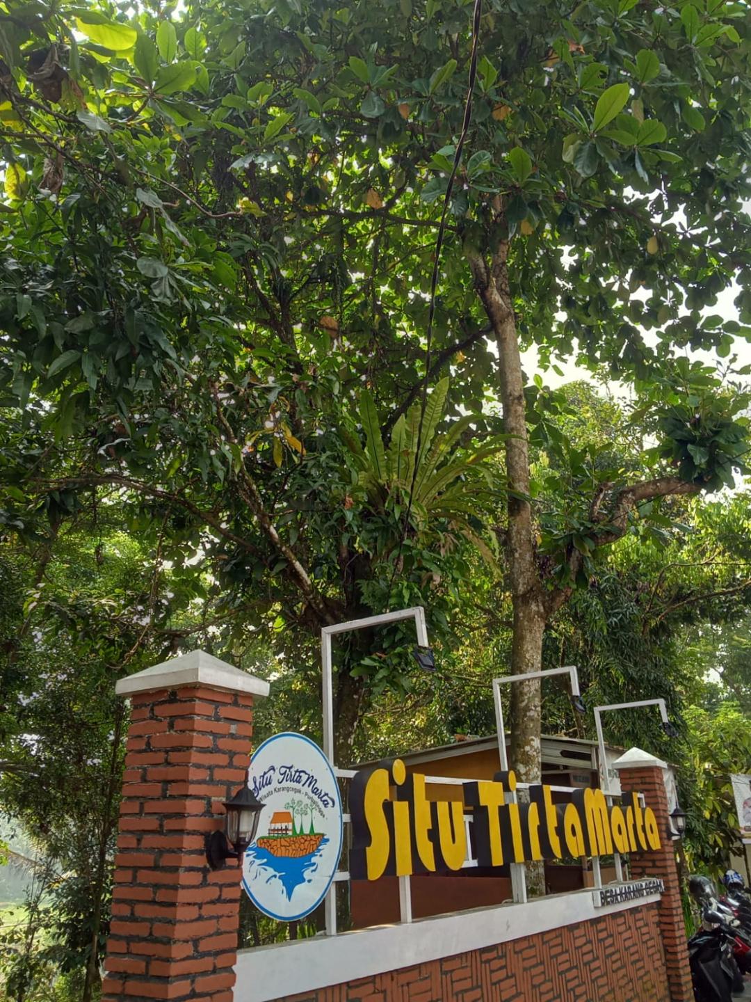 A brick-pillared park entrance under tall trees, with a hand-painted sign reading 'Situ Tirta Marta'.