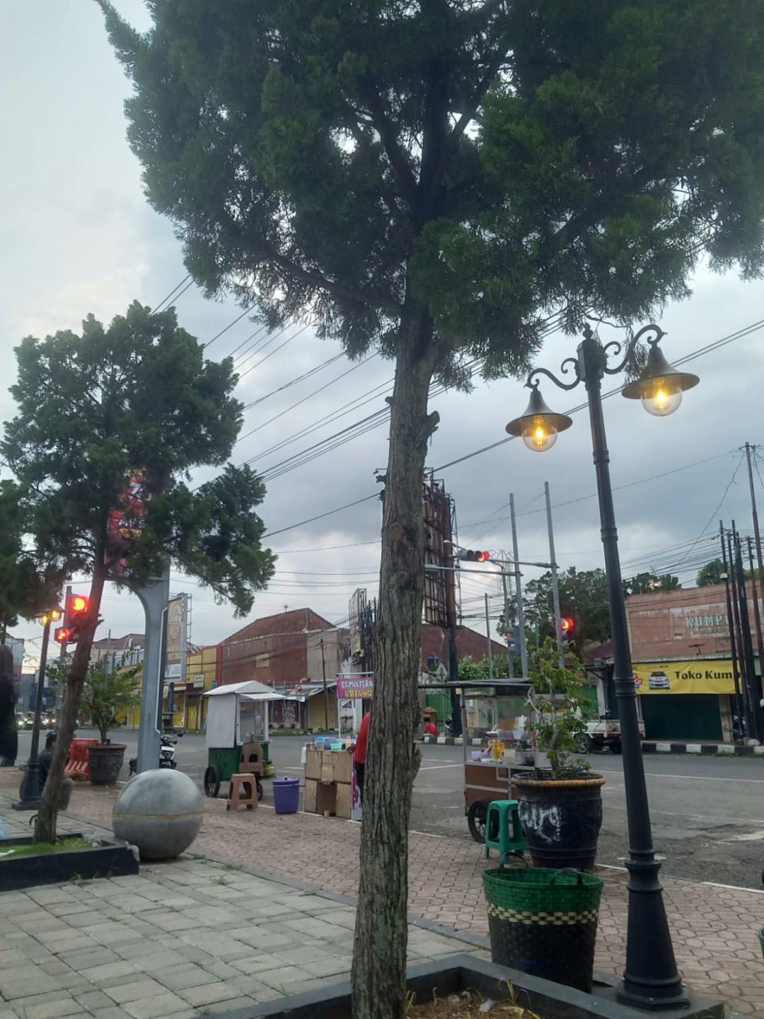 A street corner at dusk: a tall lamppost lit, a tree, a parked food cart, shops closed up beyond a quiet intersection.