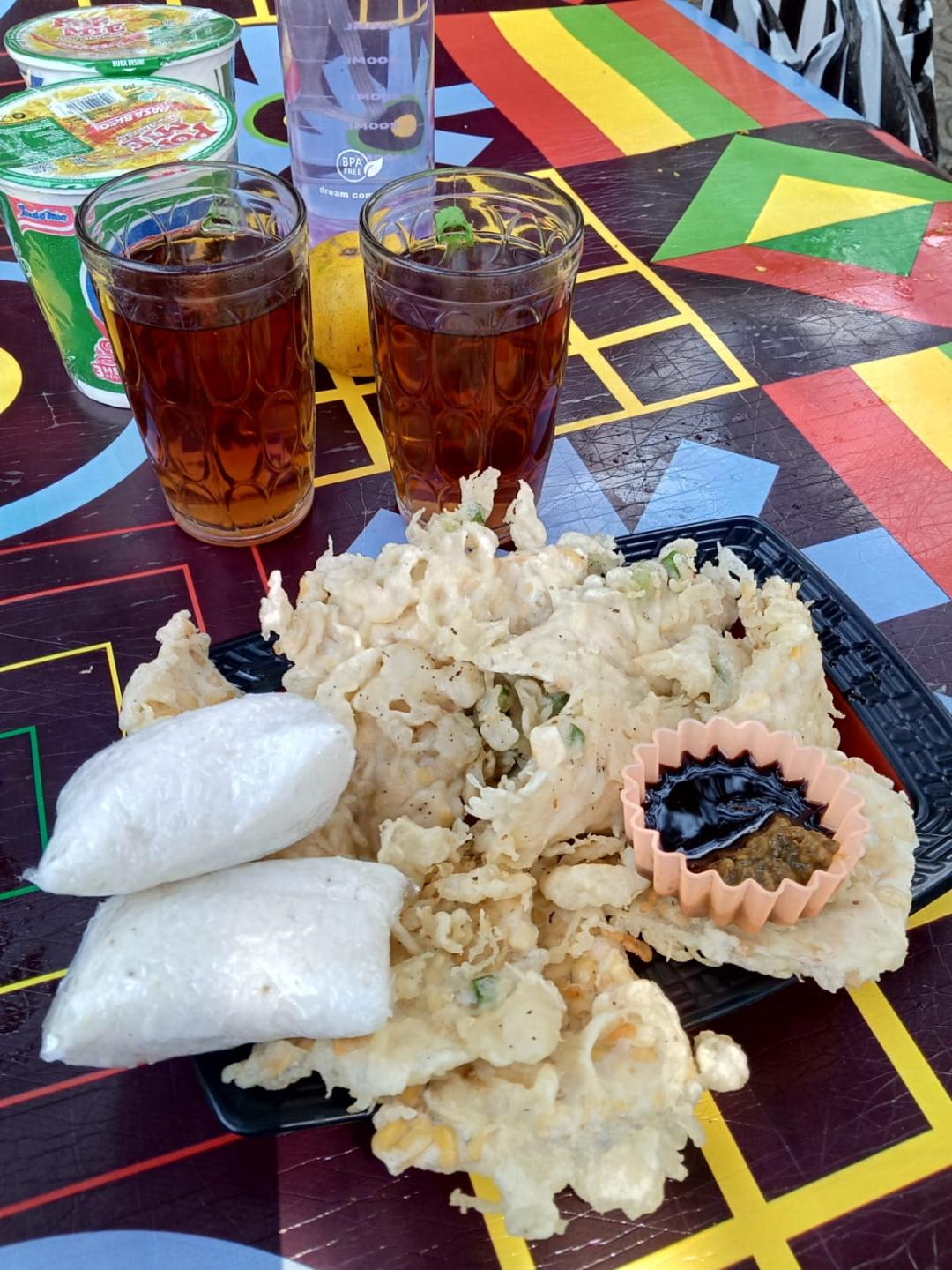 A plate of crispy bakwan fritters and rice cakes beside two tall glasses of iced tea on a brightly patterned table.