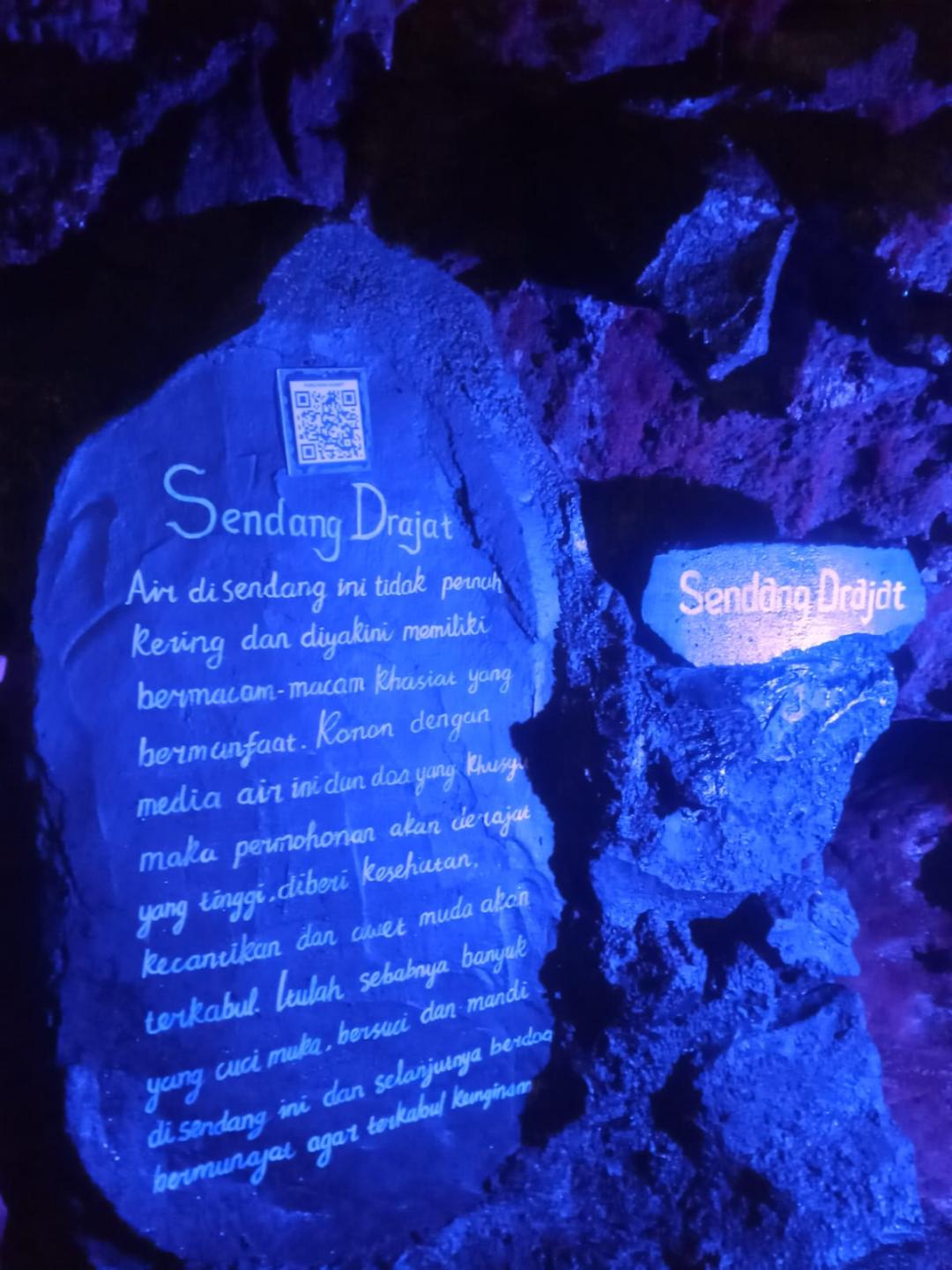 A cave wall painted blue under coloured lights, with the words 'Sendang Drajat' and a long Indonesian inscription written in white script.